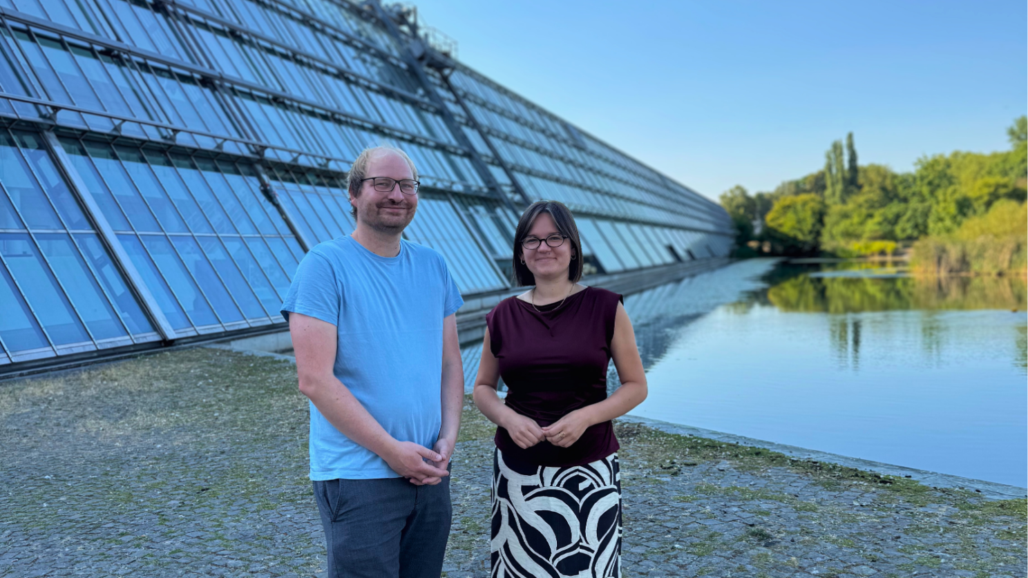 Neue Mitarbeiterin Juliane Palotz Franz Flögel (links) und Juliane Palotz stehen vor der markanten Glasfassade des Wissenschaftsparks Gelsenkirchen. Rechts erstreckt sich der Wasserteich vor dem Gebäude, im Hintergrund sind Bäume und Grünflächen zu sehen.