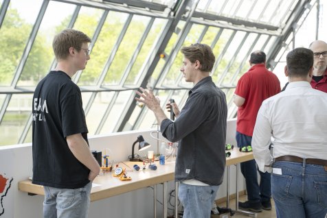 Personen stehen in einer offenen Galerie und sprechen über Experimente, die vor ihnen auf einem Tisch bereitliegen. 